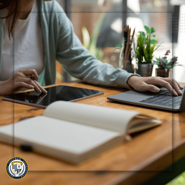 Zbliżenie na osobę pracującą równocześnie na laptopie i tablecie, z otwartą książką na biurku, obrazujące nowoczesne metody nauki kadry i płac.