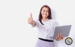 Uśmiechnięta kobieta w białej koszuli i szarej spódnicy trzyma laptop i pokazuje kciuk w górę - kadry i płace nauka to dobry wybór na rozpoczęcie kariery
