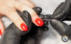 Stylistka paznokci w czarnych rękawiczkach nakładająca czerwony lakier hybrydowy na paznokcie klientki za pomocą cienkiego pędzelka, precyzyjnie aplikując produkt przy skórkach