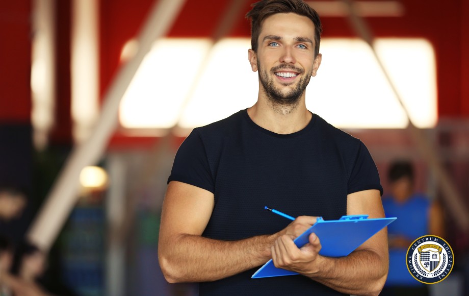 Młody, uśmiechnięty mężczyzna w czarnej koszulce z krótkim rękawem trzyma w rękach niebieski clipboard. Stoi w pomieszczeniu z czerwonymi ścianami, w tle widoczne są nieostre elementy wyposażenia. W prawym dolnym rogu znajduje się logo. Mężczyzna wygląda profesjonalnie i przyjaźnie, co sugeruje, że może być trenerem lub instruktorem