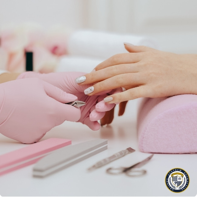 Alt: "Stylistka paznokci w różowych rękawiczkach wykonująca manicure klientce z eleganckimi paznokciami w jasnych odcieniach ze srebrnymi zdobieniami. Dłoń klientki spoczywa na różowej poduszce, a w tle widoczne są profesjonalne narzędzia do manicure