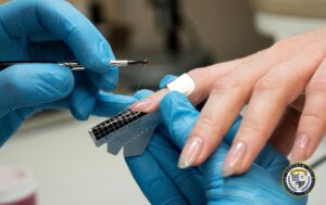 W obrazku widać technika paznokci w niebieskich rękawiczkach, który nakłada przedłużenie akrylowe na paznokieć klientki. Używa szablonu z siatką do formowania paznokcia, a dłoń klientki ma naturalne paznokcie. W prawym dolnym rogu znajduje się logo KCE.
