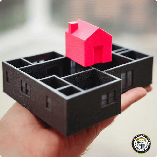 Model architektoniczny zaprojektowany w AutoCAD 3D i wydrukowany na drukarce 3D, przedstawiający czarny rzut parteru budynku z wewnętrznymi ścianami działowymi oraz małym czerwonym domkiem umieszczonym na górze, demonstrującym możliwości precyzyjnego projektowania detali.