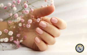 Manicure hybrydowy w delikatnym różowym odcieniu na zadbanej dłoni. Krótkie paznokcie o klasycznym kształcie z błyszczącym wykończeniem. Dłoń trzyma gałązkę gipsówki z drobnymi różowymi i białymi kwiatuszkami. Jasne tło podkreśla elegancję i naturalność stylizacji.