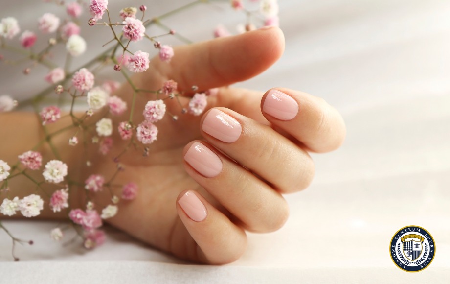 Manicure hybrydowy w delikatnym różowym odcieniu na zadbanej dłoni. Krótkie paznokcie o klasycznym kształcie z błyszczącym wykończeniem. Dłoń trzyma gałązkę gipsówki z drobnymi różowymi i białymi kwiatuszkami. Jasne tło podkreśla elegancję i naturalność stylizacji.