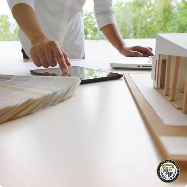 Osoba w białej koszuli pracuje przy biurku, korzystając z tabletu, laptopa i fizycznego modelu architektonicznego, analizując projekt.