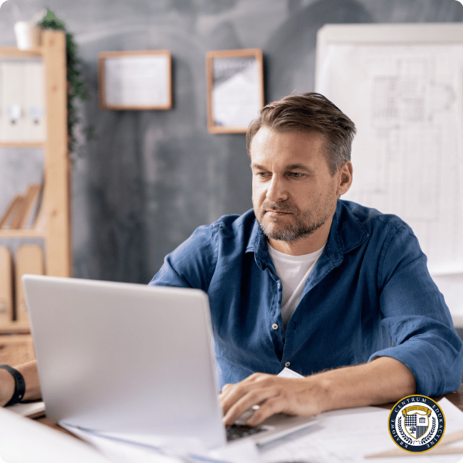 Skoncentrowany architekt w niebieskiej koszuli pracujący w programie AutoCAD 3D na laptopie w nowoczesnym biurze projektowym, z planami i szkicami widocznymi w tle, co ilustruje profesjonalne środowisko pracy podczas tworzenia zaawansowanych wizualizacji trójwymiarowych.