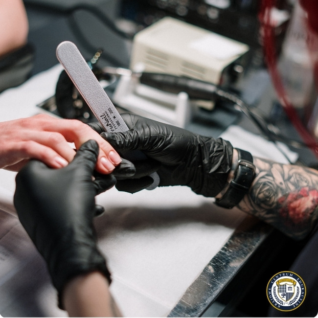 Profesjonalny manicure hybrydowy - stylistka w czarnych rękawiczkach piłuje paznokieć klientki pilnikiem o odpowiedniej gradacji, przygotowując płytkę do aplikacji lakieru hybrydowego.