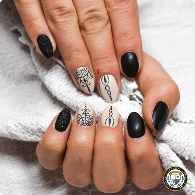 Elegancki czarno-biały manicure w stylu boho na migdałowych paznokciach. Wzór zawiera naprzemienne czarne matowe i brokatowe paznokcie oraz beżowe z czarnymi geometrycznymi wzorami i mandalami. Dłoń spoczywa na białym ręczniku. W prawym dolnym rogu znajduje się okrągłe logo.
