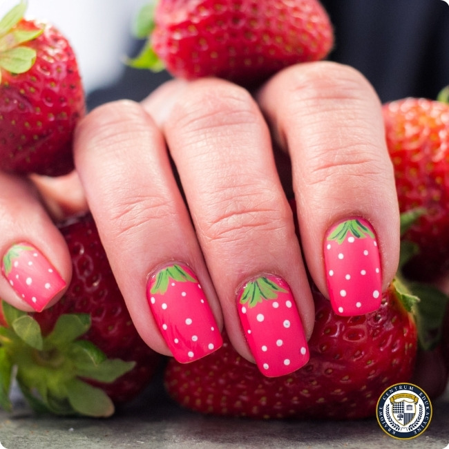 Manicure w stylu truskawkowym na tle prawdziwych truskawek. Paznokcie pomalowane są na intensywny różowy kolor z białymi kropkami, imitującymi nasiona truskawki, oraz zielonymi akcentami u nasady paznokci, przypominającymi liście. W prawym dolnym rogu znajduje się okrągłe logo
