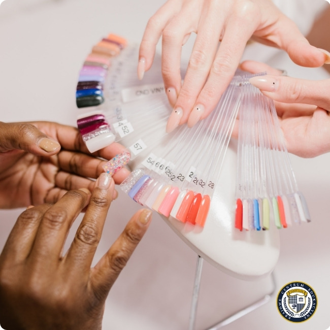 Scena z salonu manicure, gdzie dwie osoby przeglądają paletę kolorów lakierów do paznokci. Jedna dłoń trzyma przezroczysty wachlarz z próbkami kolorów, druga wskazuje na wybrany odcień. Widoczna jest szeroka gama kolorów od pastelowych po intensywne odcienie. W prawym dolnym rogu znajduje się okrągłe logo