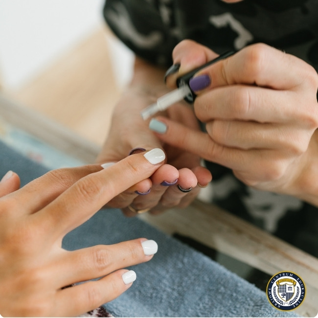 Zbliżenie na dłonie podczas wykonywania manicure, gdzie manikiurzystka z fioletowym i miętowym lakierem na paznokciach nakłada biały lakier na paznokcie klientki. Klientka ma już pomalowane na biało kilka paznokci, a zabieg odbywa się na ciemnoniebieskim ręczniku.