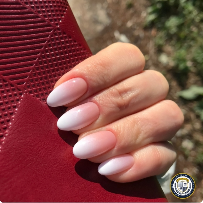 Zbliżenie na eleganckie paznokcie w kształcie migdałów z ombre manicure przechodzącym od jasnoróżowego u nasady do białego na końcówkach. Dłoń opiera się o czerwony teksturowany materiał, widoczne są cztery paznokcie.