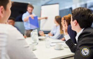 Szkolenie pracowników - grupa osób w sali konferencyjnej słucha prezentacji prowadzonej przez trenera trzymającego niebieską teczkę