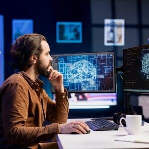 Programista studiujący wykresy i modele podczas kursu AI - kursu sztucznej inteligencji na monitorach w ciemnym biurze. Mężczyzna w brązowej koszuli pracuje w skupieniu przy stanowisku z wieloma ekranami wyświetlającymi zaawansowane wizualizacje danych