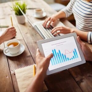 Osoba uczestniczy w kursie marketingu internetowego online, analizując na tablecie wykresy wydajności kampanii. Na drewnianym stole znajdują się filiżanki z herbatą, notatnik i przybory do pisania, podczas gdy inny uczestnik pracuje na komputerze