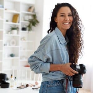 Uśmiechnięta kobieta z długimi kręconymi włosami uczestniczy w kursie na social media managera, trzymając profesjonalny aparat fotograficzny. W tle widoczne są białe regały z akcesoriami, a uczestniczka ubrana jest w jeansową koszulę