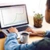 Osoba analizująca wykres wzrostu podczas kursu SEO i kursu pozycjonowania na laptopie. Na biurku widoczna jest filiżanka kawy, telefon komórkowy oraz roślina doniczkowa, co tworzy przyjazną atmosferę nauki online