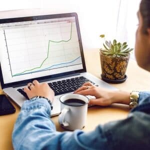 Osoba analizująca wykres wzrostu podczas kursu SEO i kursu pozycjonowania na laptopie. Na biurku widoczna jest filiżanka kawy, telefon komórkowy oraz roślina doniczkowa, co tworzy przyjazną atmosferę nauki online