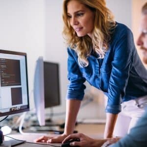 Kobieta w dżinsowej koszuli prowadzi kurs WordPress, pochylając się nad komputerem, na którego ekranie widoczny jest kod strony. Obok siedzi uczestnik szkolenia obserwujący prezentowane zagadnienia w nowoczesnym biurze