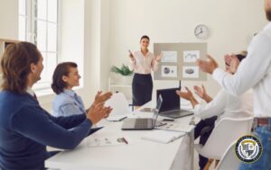 Udana prezentacja biznesowa w jasnym biurze, gdzie prezenterka pewnie odpowiada na obiekcje klientów, stojąc przy tablicy z wykresami i otrzymując pozytywne reakcje w postaci braw od uczestników spotkania. Na ścianie widoczne są wykresy biznesowe i diagramy kołowe ilustrujące dane, a na stole znajdują się otwarte laptopy i materiały do notatek