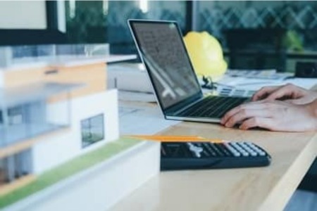 Uczestnik kursu AutoCAD online pracuje na laptopie z profesjonalnym oprogramowaniem projektowym, mając obok dokumentację techniczną i kalkulator, co wskazuje na praktyczne zastosowanie wiedzy z kursu w projektowaniu komputerowym