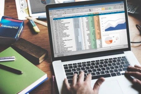Na laptopie wyświetlony jest arkusz Excel z kursem tabele przestawne, pokazujący rozbudowane zestawienia danych, wykresy i analizy. Na drewnianym biurku obok komputera znajdują się kolorowe notatniki i przybory do pisania