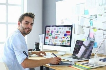 Młody grafik uczestniczy w kursie grafiki komputerowej online, pracując nad projektem na profesjonalnym stanowisku wyposażonym w komputer stacjonarny i laptop, otoczony narzędziami projektowymi i tablicą inspiracji w jasnym, nowoczesnym biurze