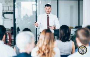 Profesjonalny trener prowadzi szkolenie marketingowe dla grupy uczestników w nowoczesnej sali konferencyjnej. Mężczyzna w białej koszuli i bordowym krawacie gestykuluje, trzymając tablet, podczas gdy zróżnicowana grupa słuchaczy skupiona jest na jego prezentacji. Jasne, szklane ściany i minimalistyczny wystrój tworzą profesjonalną atmosferę sprzyjającą nauce