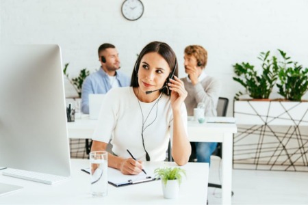 Młoda kobieta w słuchawkach podczas szkolenia z telemarketingu pracuje przy komputerze i robi notatki. W tle widoczni są inni uczestnicy w jasnym, nowoczesnym biurze z zielonymi roślinami