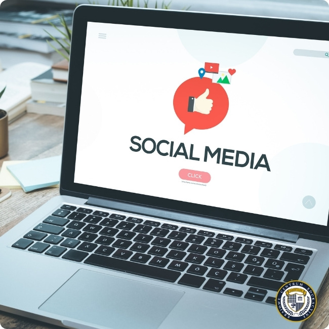 Laptop na drewnianym biurku wyświetlający logo 'SOCIAL MEDIA' z czerwoną ikoną kciuka w górę oraz symbolami mediów społecznościowych. W tle widoczne materiały biurowe i roślina