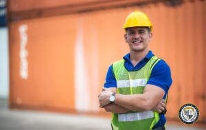 Uśmiechnięty pracownik w odzieży ochronnej BHP stoi na tle pomarańczowego kontenera. Ma na sobie żółty kask ochronny, niebieską koszulkę polo oraz odblaskową kamizelkę w żółto-białych kolorach. Jego pozycja ze skrzyżowanymi rękami i pewna postawa sugerują profesjonalizm i doświadczenie w dziedzinie bezpieczeństwa pracy