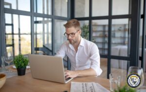 Młody biznesmen w białej koszuli i okularach pracuje przy laptopie w nowoczesnym biurze z przeszklonymi ścianami. Na drewnianym biurku znajduje się gazeta, szklanka wody i roślina doniczkowa. Zdjęcie ilustruje artykuł sponsorowany.