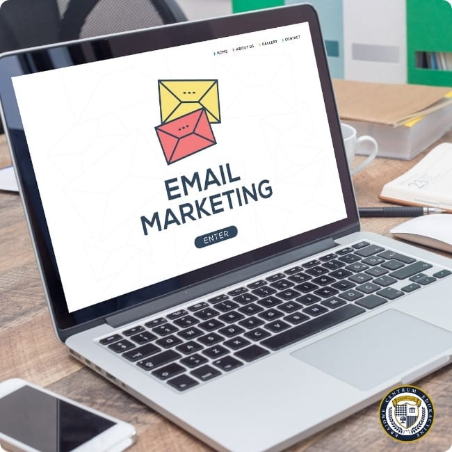 Laptop na drewnianym biurku wyświetlający logo email marketingu z ikonami żółtej i różowej koperty oraz przyciskiem 'ENTER'