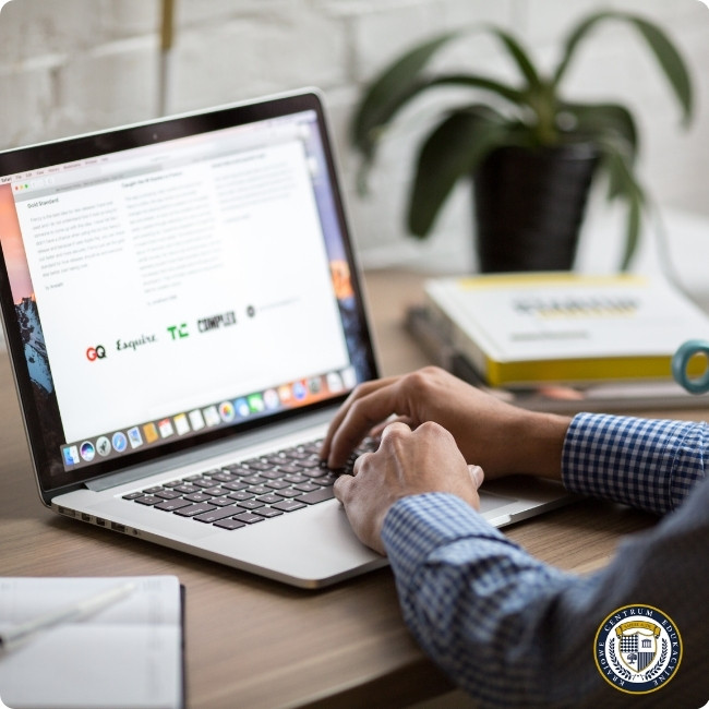 Zdjęcie przedstawia osobę w koszuli w niebieską kratę, piszącą na laptopie MacBook ustawionym na drewnianym biurku. Na ekranie widoczny jest dokument tekstowy oraz kolorowe logo. Obok laptopa znajduje się roślina doniczkowa i żółta książka. W tle widać białą ścianę z cegły. W prawym dolnym rogu znajduje się okrągłe logo KCE.