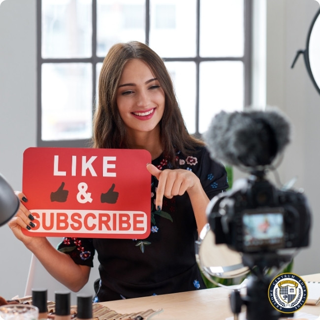 Młoda content creatorka nagrywająca wideo do mediów społecznościowych, trzymająca czerwoną tabliczkę z napisem 'LIKE & SUBSCRIBE'. W kadrze widoczna profesjonalna kamera, przedstawiająca kulisy tworzenia treści dla mediów społecznościowych