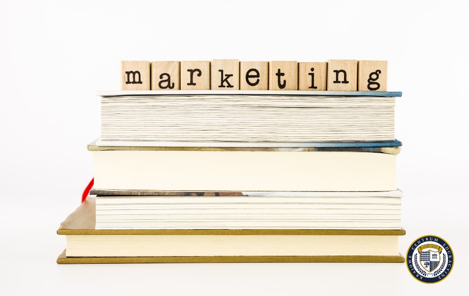 Zdjęcie przedstawia stos książek, na których ułożono drewniane literki układające się w słowo "marketing". Książki są różnej grubości, w kolorach białym, kremowym i złotym. W tle widać również logo organizacji. Obraz symbolizuje zasoby wiedzy o marketingu - najlepsze książki o marketingu zgromadzone w jednym miejscu.