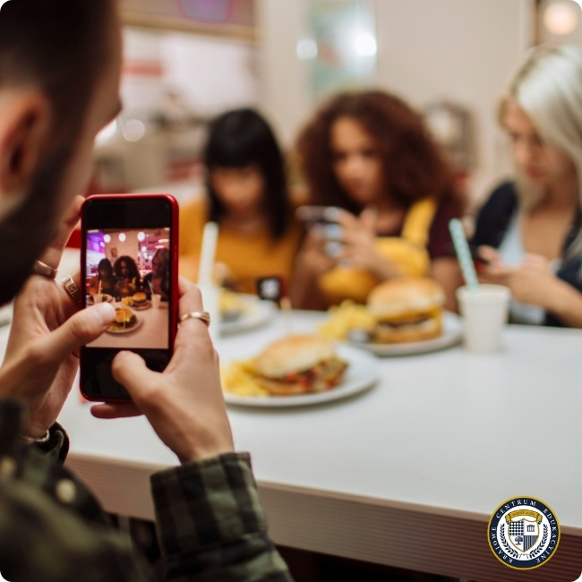 Osoba fotografująca posiłek i przyjaciół do mediów społecznościowych - w kadrze widoczny smartfon oraz grupa osób przy stole z hamburgerami i napojami, ukazując typową sytuację dzielenia się momentami w social mediach