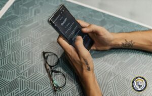 Dłonie z tatuażami trzymające smartfon z otwartą aplikacją ChatGPT, obok leżą okulary na geometrycznym blacie - poradnik jak korzystać z ChatGPT