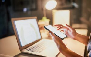 Osoba sprawdzająca social media marketing na smartfonie, pracując jednocześnie na laptopie w wieczornym oświetleniu biurowym