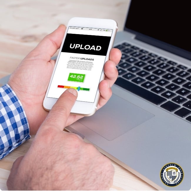 Na zdjęciu widać dłoń osoby trzymającej biały smartfon, na którym wyświetla się aplikacja lub strona internetowa z napisem "UPLOAD" na czarnym tle. Poniżej widoczny jest tekst "FASTER UPLOADS" oraz kolorowy wskaźnik prędkości pokazujący wartość 42,62. Osoba ma na sobie koszulę w niebieską kratkę i obsługuje telefon, znajdując się przy laptopie, którego część klawiatury jest widoczna w tle. Całość znajduje się na jasnej drewnianej powierzchni.