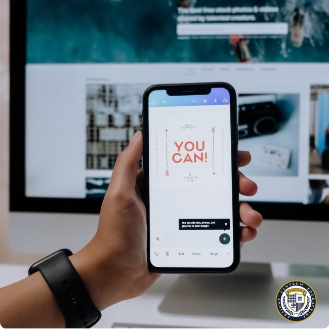 Dłoń trzymająca smartfon z motywacyjnym komunikatem 'YOU CAN!' na ekranie, w tle widoczny monitor komputera