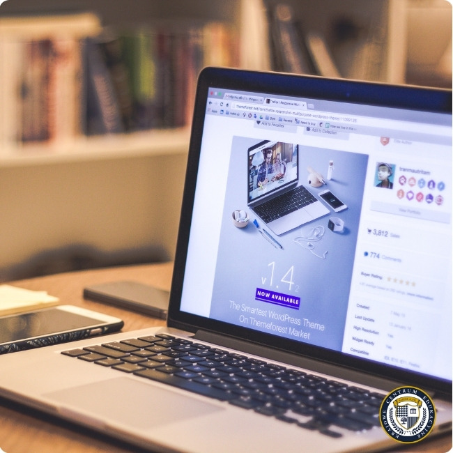 Laptop na biurku wyświetlający reklamę motywu WordPress w wersji 1.4, w tle widoczne półki z książkami.