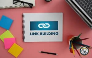 Notes z napisem 'Link Building' na biurku z różowawym tłem, otoczony kolorowymi karteczkami, długopisem, okularami, kubkiem z kredkami i laptopem – koncepcja strategii link building w marketingu internetowym