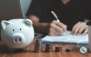 Osoba wypełniająca dokumenty przy biurku ze skarbonką i monetami - ilustracja procesu ubiegania się o dofinansowanie do szkolenia