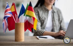Flagi europejskich krajów w drewnianym pojemniku na biurku, symbolizujące zatrudnienie cudzoziemca w Polsce. W tle osoba pracująca przy laptopie.
