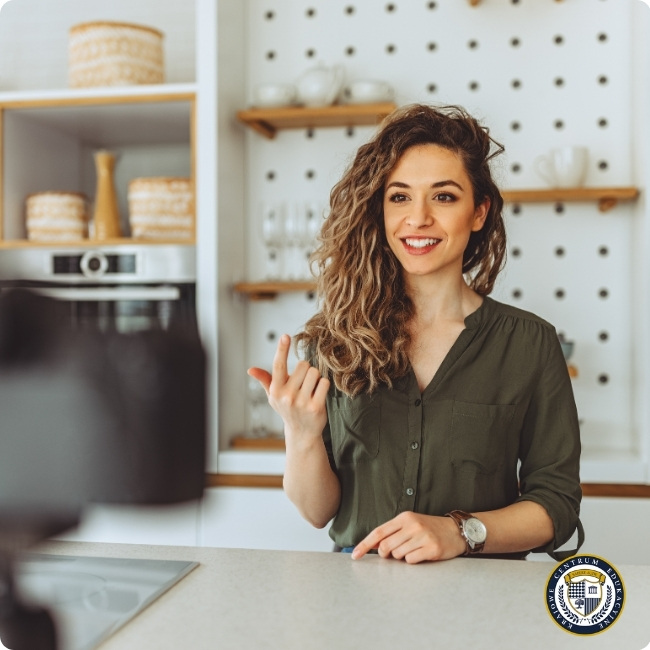 Uśmiechnięta kobieta w oliwkowej koszuli podczas nagrywania wideo o kreowaniu wizerunku w jasnej kuchni z drewnianymi półkami - przykład profesjonalnej autoprezentacji.