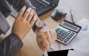 Osoba sprawdzająca zegarek przy biurku z laptopem i kalkulatorem