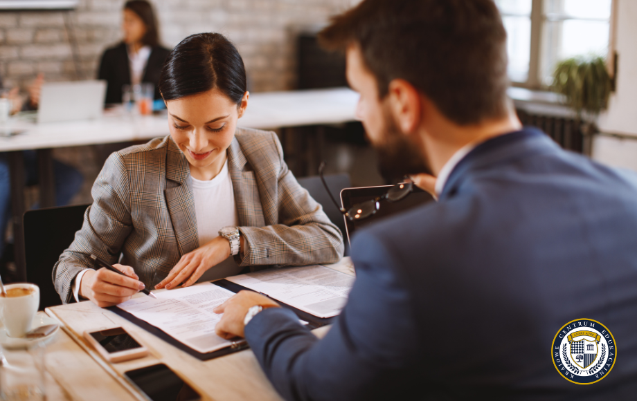 Kobieta w eleganckim żakiecie podpisuje dokument (prawdopodobnie umowę o pracę) leżący na stole, podczas gdy mężczyzna w garniturze (być może pracodawca lub rekruter) wskazuje coś palcem na tym dokumencie.