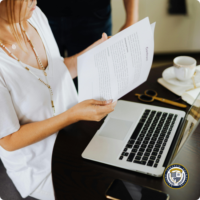 Zbliżenie na kobietę w białej koszulce, która trzyma i uważnie czyta dokument (prawdopodobnie umowę) przed laptopem.